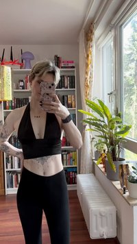 Juoda viršūnė su apykakle ir giliu iškirpimu, derinama su juodomis kelnėmis su aukštu juosmeniu. Kūną puošia tatuiruotės, o fone matoma augalas.