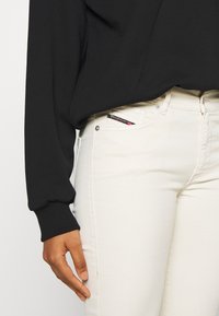 Sudadera negra con puños acanalados combinada con jeans de mezclilla blancos, que presentan un acento de bolsillo y detalles de costura en contraste.