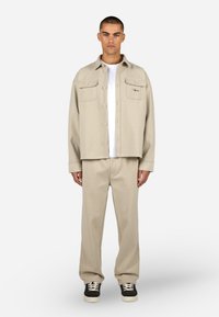 Beige dzseki két mellzsebellel és gombokkal, párosítva azonos színű bézs nadrággal. A modellen fehér póló és fekete cipő van.