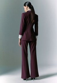 Completo bordeaux con giacca su misura dal colletto affilato e pantaloni svasati. Polsini della camicia bianca visibili, texture liscia e tacchi lucidi.