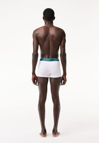 Boxerii alba cu o talie teal care prezintă logo-ul Lacoste, expuși din spate pe un model cu picioare goale, pe un fundal deschis.