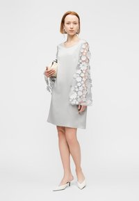 Φόρεμα από γκρι σατέν με μακριά διαφανή μανίκια διακοσμημένα με λευκά ανθισμένα εφαρμοστά, συνδυασμένο με λευκές μυτερές γόβες και μικρή τσάντα χειρός.