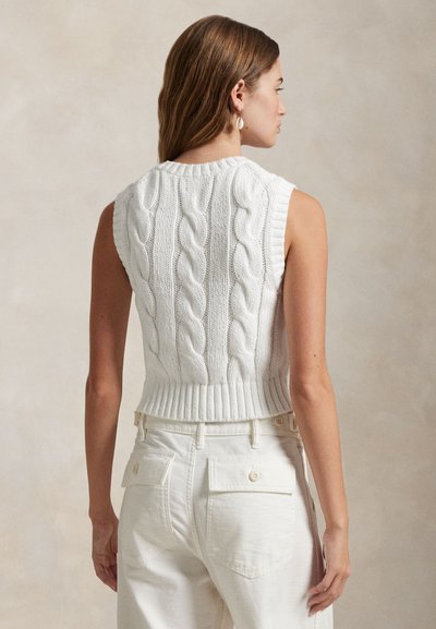 Balta adīta piedurkņu džemperis bez piedurknēm ar ciešu riboto apakšu. Valkāts kopā ar augstās jostasvietas krēmkrāsas biksēm, kas izceļ teksturētu rakstu.