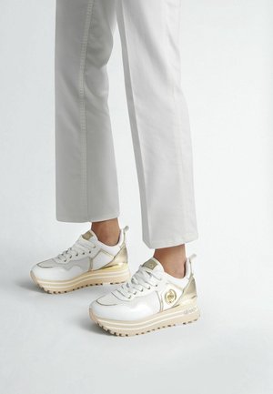 Osoba nosząca białe i złote sneakersy na platformie w połączeniu z białymi spodniami, stojąca na jednolitym jasnym tle.