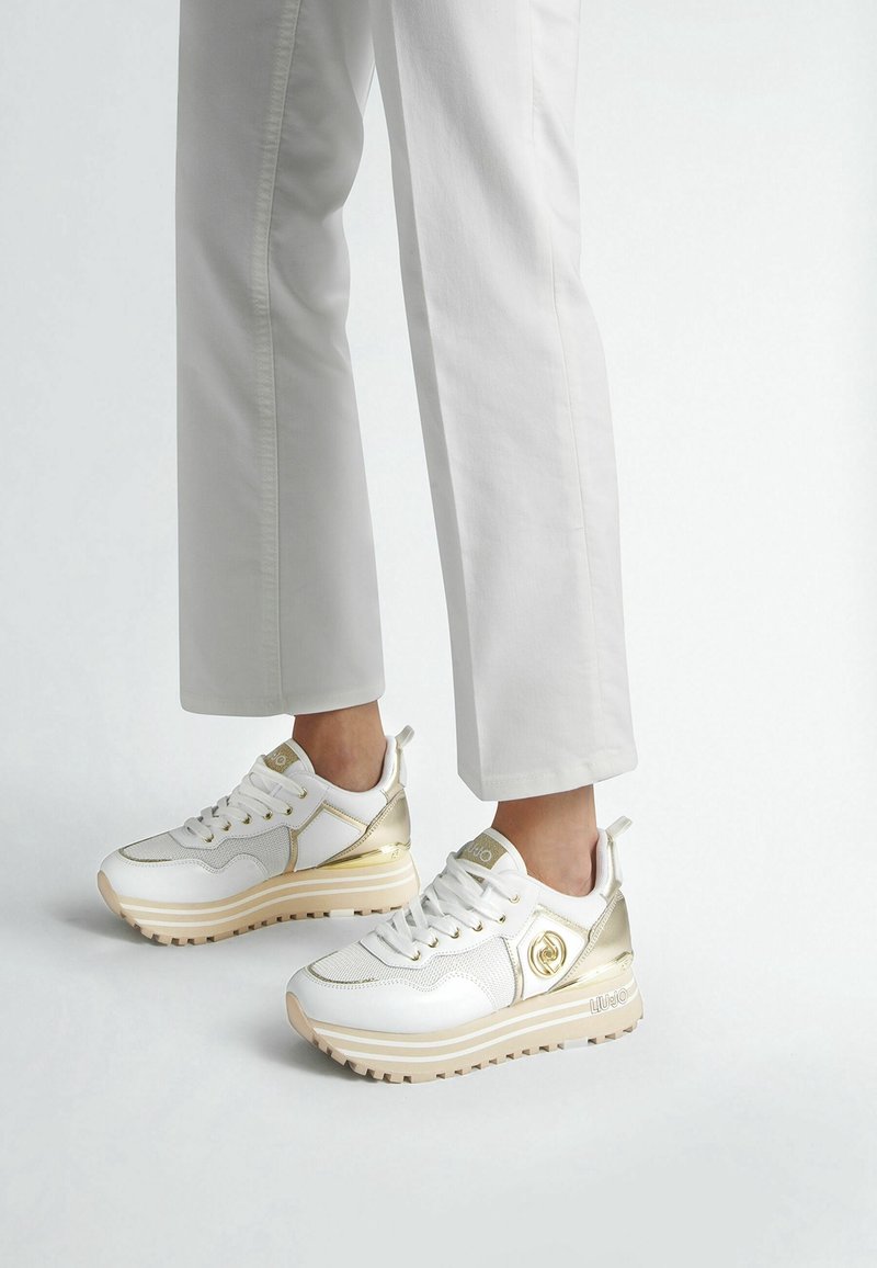 Personne portant des baskets compensées blanches et dorées assorties à un pantalon blanc, debout devant un fond clair uni.