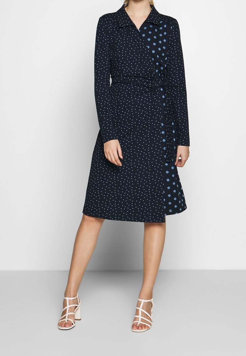 Femme portant une robe portefeuille bleu marine mi-longue avec des motifs de pois bleu clair mélangés et des sandales blanches à talons avec brides.