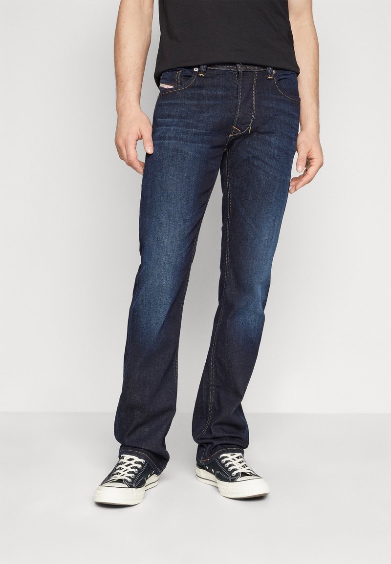 Jeans en denim bleu foncé avec une coupe droite, présentant des coutures contrastées et un design à cinq poches. Portés avec des baskets noires.