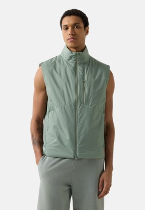 Junger Mann mit kurzem lockigem Haar, der eine hellgrüne ärmellose Weste mit Reißverschluss und passende Hose trägt, und mit den Händen in den Taschen steht.