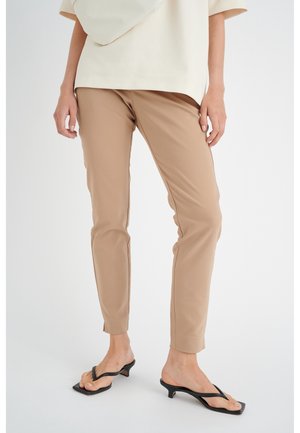 Woman wearing beige fitted pants, cream top, black heeled thong sandals, standing against a white background.