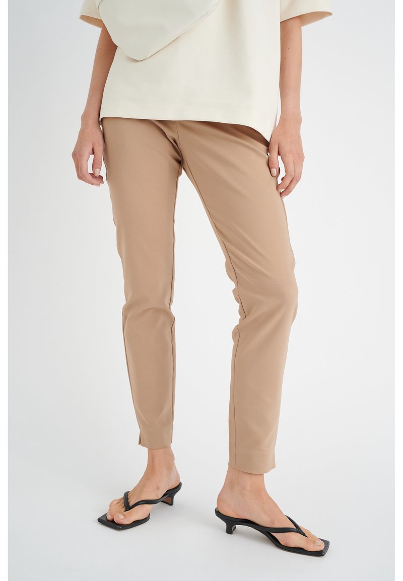 Femme portant un pantalon beige ajusté, un haut crème, des sandales à talons noires, se tenant contre un fond blanc.