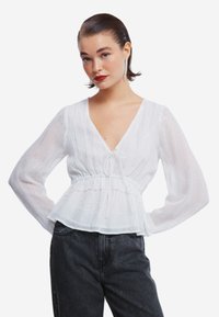 Hvid, gennemsigtig bluse med lange ærmer, v-hals og snørre i taljen. Har diskrete strukturerede striber og lagdelte flæser i kanten.