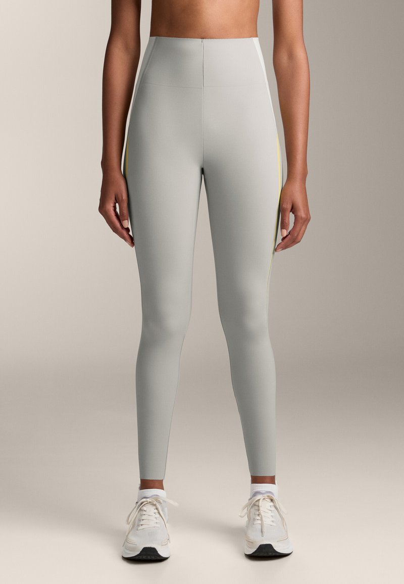Világosszürke leggings magas derékkal, sima textúrával és sárga oldalsávokkal; fehér sportcipővel párosítva.