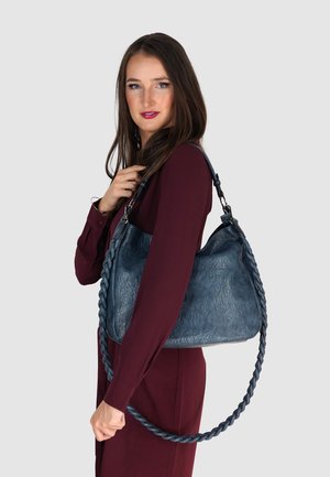 Blaue, texturierte Leder-Umhängetasche mit einem geflochtenen Trageriemen, die einen geräumigen Körper und ein schlankes Design aufweist, gehalten von einem Modell in einem burgunderfarbenen Kleid.