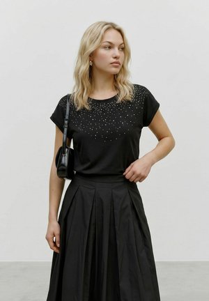 Femme aux cheveux blonds portant un t-shirt noir clouté, une jupe plissée noire et un petit sac à épaule noir, regardant sur le côté.