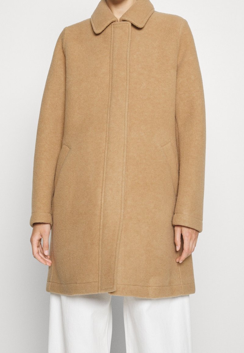Beige wool coat with a structured collar, featuring side pockets and visible stitching details. The coat has a mid-length cut and a soft texture.