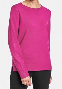 Osoba ubrana w długi rękaw, jasnoróżowy sweter z okrągłym dekoltem i bocznymi rozcięciami, w połączeniu z czarnymi spodniami, na jednolitym tle.