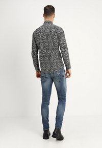 Uomo di spalle che indossa una camicia a maniche lunghe a fiori bianca e nera, jeans skinny strappati sbiaditi e stivali alla caviglia neri.