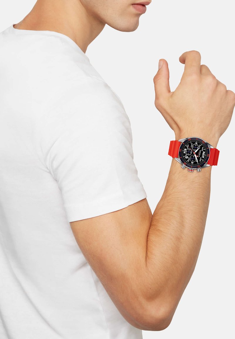 Montre noire avec des caractéristiques de chronographe, materiel en argent et un bracelet en silicone rouge vif, portée sur un poignet, complétée par un T-shirt blanc.