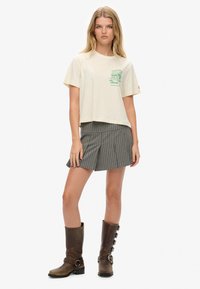Beige cropped t-shirt med grøn grafisk tekst. Stribede højtaljede shorts i grå. Brun knælang støvler med spænder. Ensartet baggrund.
