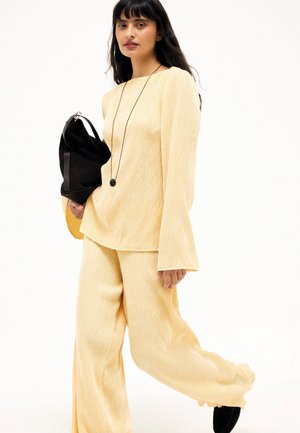 Woman with long dark hair wearing light yellow textured long-sleeve top and wide-leg pants, holding a black bag and wearing a long necklace.