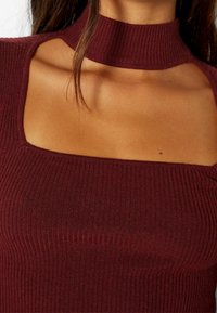 Top de punto acanalado burdeos con escote cuadrado y cuello alto, que presenta detalles con cortes en los hombros. Textura suave y diseño ajustado.