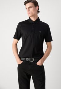 Black polo shirt in smooth fabric with a two-button placket and a logo patch on the chest, paired with black textured jeans and a leather belt.