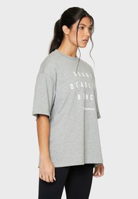 Harmaa isokokoinen t-paita, jossa on lyhyet hihat. Edessä on rohkea valkoinen teksti: "SQUAT DEADLIFT BENCH" ja "SQUATPROOF" -logo alhaalla.
