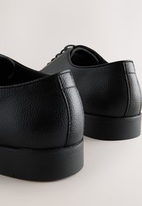 Schwarze Lederschuhe mit genarbter Textur, flachem Design, abgerundeten Zehen und flachen Gummisohlen. Schnürsenkel sind vorne sichtbar.