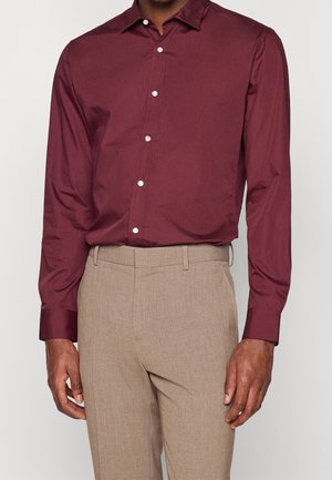 Uomo con camicia bordeaux a maniche lunghe abbottonata infilata in pantaloni beige su misura, in piedi con le braccia rilassate.