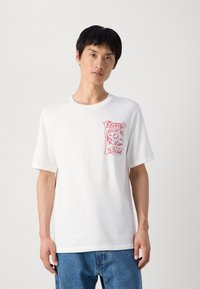 Hvid bomulds t-shirt med rund hals, der har et rødt grafisk design på venstre brystområde. Korte ærmer, afslappet pasform. Denim jeans.