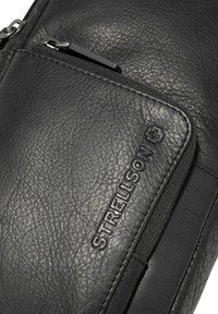 Bolso de cuero negro con superficie texturizada, compartimentos con cremallera y el logo grabado "Strellson" en la parte delantera.