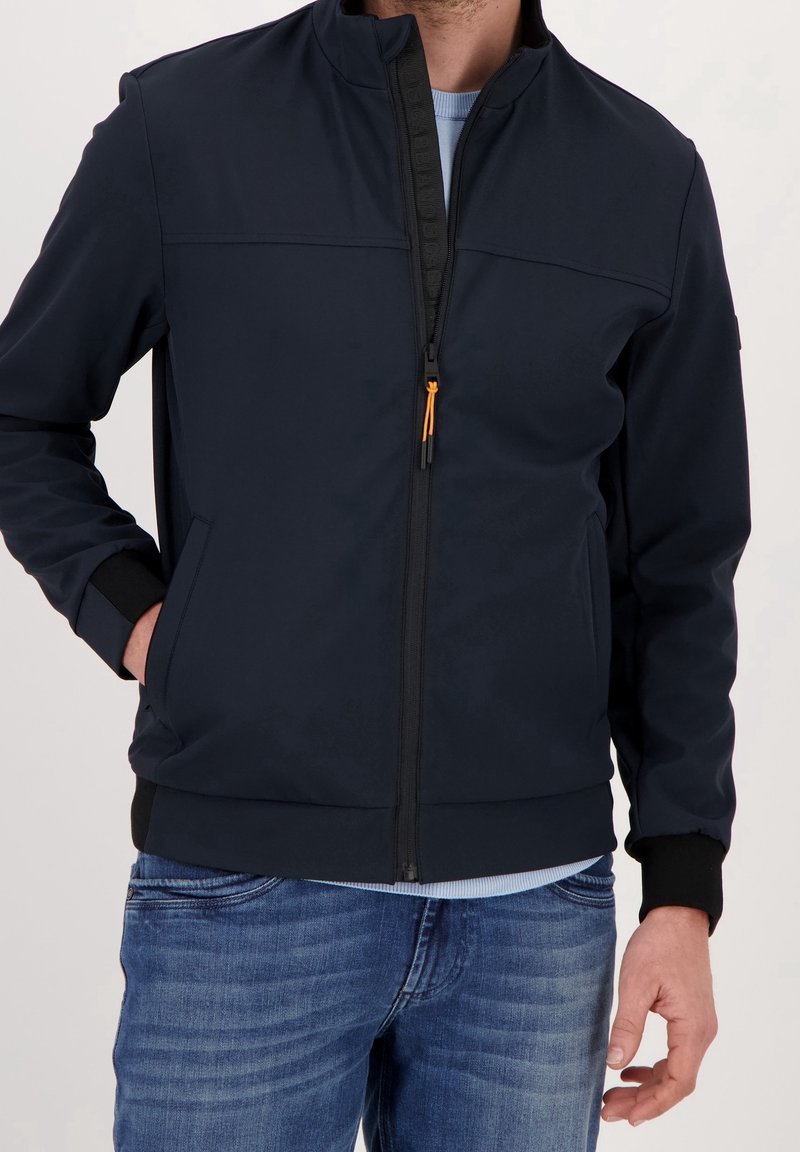 Homme portant une veste noire zippée avec poignets et col côtelés, jean bleu et chemise bleu clair en dessous, main dans la poche.
