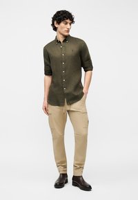 Νεαρός άνδρας με πουκάμισο σε ελιά πράσινο, μπεζ παντελόνι cargo και σκούρα καφέ μπότες, στέκεται με το ένα χέρι στην τσέπη.