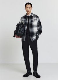 Chemise à carreaux noir et blanc, pull noir, pantalon noir et bottines noires. Tient un sac noir texturé avec une bandoulière.