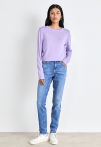 Camisola roxa de mangas longas combinada com jeans de brim azul claro e sapatilhas brancas. A camisola tem um decote redondo e mangas justas.