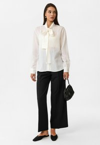 Witte, gestreepte blouse met een grote strik bij de hals, gecombineerd met zwarte wijdvallende broek en een gestructureerde zwarte handtas. Zwarte platte schoenen.