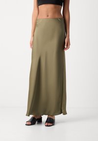 Femme portant une jupe maxi en satin olive, un crop top noir et des sandales à talons blocs ouvertes noires, se tenant devant un fond blanc uni.