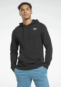 Sudadera de algodón negra con un bolsillo frontal, capucha con cordón, y logo blanco de Reebok en el pecho. Combinada con pantalones de chándal azules.