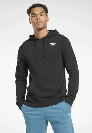 Sudadera de algodón negra con un bolsillo frontal, capucha con cordón, y logo blanco de Reebok en el pecho. Combinada con pantalones de chándal azules.