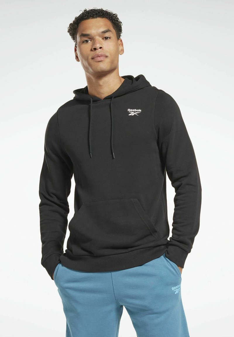 Sudadera de algodón negra con un bolsillo frontal, capucha con cordón, y logo blanco de Reebok en el pecho. Combinada con pantalones de chándal azules.