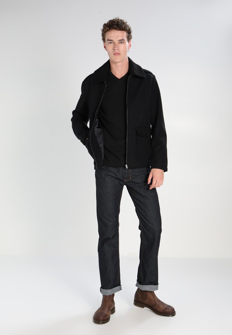 Svart zip-up jacka med en texturerad krage, bärs över en svart skjorta, matchad med mörka denimjeans och bruna läderstövlar.