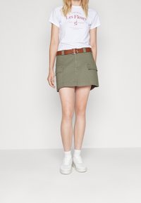 Jupe cargo kaki avec deux poches avant, associée à un t-shirt blanc avec un graphisme rouge, et des baskets blanches à semelle épaisse.