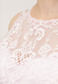 Krajkový top s detailním květinovým výšivem v bílé barvě na průsvitné světle růžové tkanině, s kulatým výstřihem a jemným zpracováním.