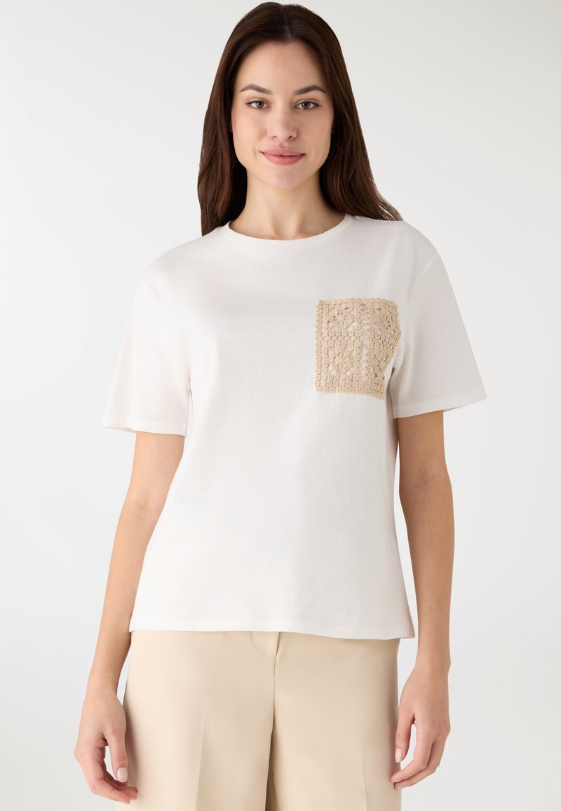 Kremowy T-shirt z kwadratową beżową koronkową kieszonką. Krótkie rękawy, miękka bawełniana tkanina, luźny krój, gładka faktura, minimalistyczny design.