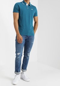 Teallin polo-paita, jossa on valkoiset kaulustiedot, yhdistettyinä keskivärisiin kuluneisiin farkkuihin ja valkoisiin tennareihin. Yksinkertainen muotoilu, lyhyet hihat.