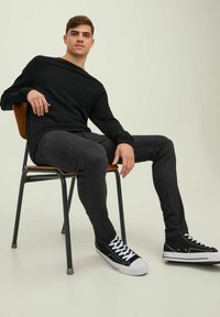 Svart langermet skjorte, slim-fit mørkejeans og svarte høy-top sneakers med hvite såler, sittende på en trestol mot en lys bakgrunn.