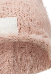 Stickad rosa mössa gjord av fluffigt garn, med en vikbar brätte och en tygmärkning med präglad logotyp. Mjuk och texturerad framtoning.