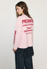 Rózsaszín hosszú ujjú ing, piros felirattal a háton: "PICANTE LIKE CHILIS" és "Delicioso" az ujjon; laza szabású és puha anyagból készült.