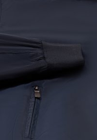 Maniche di giacca navale con polsino a coste e tasca con zip su sfondo di tessuto liscio.