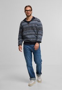 Navy- und hellblau gemusterter Pullover mit Viertelreißverschluss, mit gerippten Bündchen, getragen über einem schwarzen Shirt, kombiniert mit blauen Jeans und Turnschuhen.